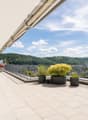 Balcony with white tiled floor, metal railing, potted plants, and a scenic view of a green, hilly landscape.