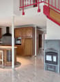 Open-plan kitchen and living area with wooden cabinets, a central island with bar stools, a modern range hood, and a fireplace with a firewood rack nearby.