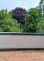 A flat roof terrace with a tiled surface and a low concrete parapet wall, surrounded by lush green trees and a distant view of a spire.