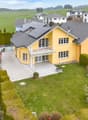 Aerial view of a two-story yellow house with a grey roof, surrounded by a well-maintained lawn, garden, and neighboring houses in a suburban setting.