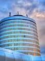 A modern, cylindrical skyscraper with reflective glass windows and a rounded top, featuring communication antennas on the roof.