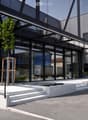 Building. Modern building with large glass windows and a sleek, contemporary design. Features a raised ground floor, small water feature, and small landscaped area with plants and trees.