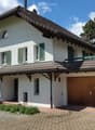 House. Two-story white house with green shutters, wooden garage, and solar panels on the roof. The house features a balcony on the upper level and is surrounded by greenery.