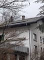 House. A two-story house with white walls, multiple windows, and a tiled roof, partially obscured by trees and branches.
