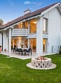 House. Modern two-story house with white exterior, large windows, and a spacious garden featuring a dining set, fire pit, and well-maintained lawn.