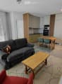 Furnished Room. Open-plan living and dining area with a grey sofa, wooden coffee table, dining table with chairs, and a small kitchen area with cabinets and appliances.