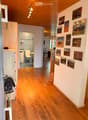 Corridor / Hallway. A well-lit hallway with wooden flooring, white walls, and several framed pictures. A small bathroom is visible to the left, and a dining area is visible to the right.