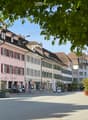 Garden / Outdoor Area. A European town square with colorful buildings, outdoor seating, and trees. The square is paved and features potted plants and umbrellas.