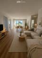 Modern living and dining area with a sectional sofa, coffee table, dining table with chairs, and a TV stand. The space features wooden flooring, large windows with sheer curtains, and decorative plants.