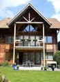 House. Two-story wooden house with a triangular roof, large windows, and a balcony. The house is surrounded by a well-maintained garden with a patio area, outdoor furniture, and potted plants.