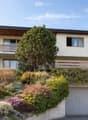Two-story house with a raised ground floor, modern design, large windows, and a balcony on the upper level. The house features a garage and is surrounded by well-maintained gardens and trees.