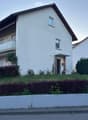 Two-story white house with a pitched roof, balcony, and small garden area.