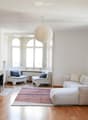 Bright living room with white walls, wooden flooring, large windows, a fireplace, a white sectional sofa, two white armchairs, a small wooden table, a floor lamp, a pendant lamp, and a potted plant.