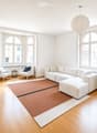 Bright living room with large arched windows, white walls, light wood flooring, a sectional sofa with an ottoman, two armchairs, a small side table, a floor lamp, a potted plant, and a picture frame on the wall.