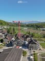 Aerial view of a rural village with traditional houses, a church, and a construction site with a crane in the background, surrounded by green fields and mountains.