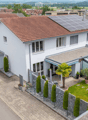 House. A two-story white house with a red-tiled roof, solar panels on the roof, and a modern garage. The house features large windows and a well-maintained garden with a palm tree, shrubs, and a small lawn.