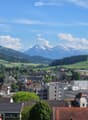 A panoramic view of a town with a mix of residential and commercial buildings, surrounded by green fields and mountains in the background.