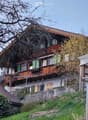 Two-story wooden house with green shutters, wooden balcony, and covered patio area.