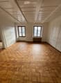 Empty room with wooden herringbone flooring, white walls, and two windows with brown frames. The ceiling has exposed wooden beams.