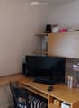 A small, furnished room with a wooden table, a black chair, a large screen, a remote control, a small cabinet, and various items on the shelves and table.