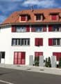 House. Two-story house with white walls, red shutters, and a red-tiled roof. Features multiple windows, a front door, and small stone planters with greenery.