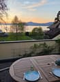 Balcony with a wooden table set for two, overlooking a lake and mountains at sunset.