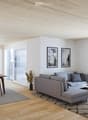 Furnished Room. Modern living room with a grey sectional sofa, coffee table, dining table with chairs, kitchen area with oven and fridge, and large windows with a view of the garden.