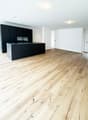 Empty room with light wooden flooring and white walls, featuring a dark kitchen island with cabinets and a sink.