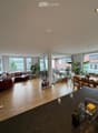 Open-plan living and dining area with wooden flooring, large windows, and a balcony. Features a red sofa, dining table with chairs, and a kitchen counter with a faucet and various items.