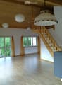 Bright, empty room with wooden flooring, large windows with yellow curtains, wooden staircase leading to an upper level, and several hanging lamps.