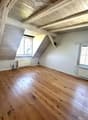 Empty attic room with wooden floors, white walls, exposed wooden beams, and two windows with blinds.
