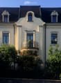 A three-story white house with ornate architectural details, including decorative window surrounds and a small balcony with a potted plant. The house features multiple windows and is surrounded by lush greenery and flowering bushes.