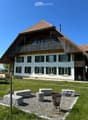 House. Traditional two-story house with white walls, dark green shutters, and a wooden balcony on the upper floor. The roof is red-tiled with a prominent wooden overhang. The surrounding area includes a lawn, a small seating area with stone blocks and a m