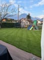 Well-maintained lawn with a wooden deck, covered grill, and a children's play area including a slide and swing set. The background features a hedge, trees, and distant mountains.
