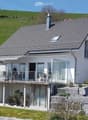 A modern two-story house with a grey tiled roof, white exterior, and a spacious balcony with glass railings. The house is surrounded by a well-maintained garden with potted plants and a stone wall.