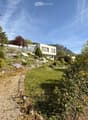 A modern house with a raised ground floor, surrounded by a landscaped garden with a gravel path, lush greenery, and a scenic mountain view in the background.