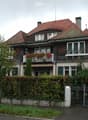 Two-story house with red-tiled roof, multiple windows, balconies, and surrounding greenery.