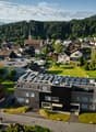 Aerial view of a modern building with solar panels on the roof, surrounded by a small garden and integrated into a village setting with traditional houses and green spaces.