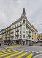 A multi-story building with a distinctive tower and spire, located at a street corner with a pedestrian crossing.