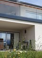 Modern two-story house with a large glass balcony on the upper level, a covered patio area with a grill, and a well-maintained garden.