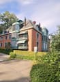 A multi-story brick house with green balconies, multiple windows, and a well-maintained hedge in front.