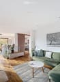 Modern living room with a green sectional sofa, white coffee table, fireplace, artwork, and large windows.