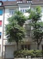 6 story white apartment building with trees and bushes in front, white garage door, red awnings, brown facade on the right side, graffiti on the right building