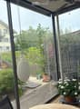 A well-maintained garden area with wooden decking, potted plants, a small tree, and a pink umbrella. The space is enclosed by glass walls, with a view of neighboring houses and greenery.