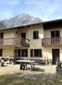 Two-story house with beige walls, wooden shutters, and balconies overlooking a scenic mountain view. The yard includes a table, chairs, and various tools and items.