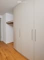 A narrow hallway with light wooden flooring, white walls, built-in white cabinets with silver handles, and a closed door at the end.