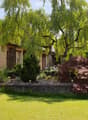 Single-story house with beige exterior, surrounded by a well-maintained garden with trees, shrubs, and a lawn.
