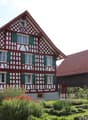 Traditional half-timbered house with red and white exterior, green shutters, and a well-maintained garden with various plants and shrubs.