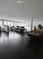 Modern office space with black flooring, large windows, office desks, chairs, a sofa, and plants.