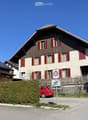 Two-story house with red shutters, solar panels on the roof, and a small balcony. A red car is parked in front, and there are speed limit signs visible.